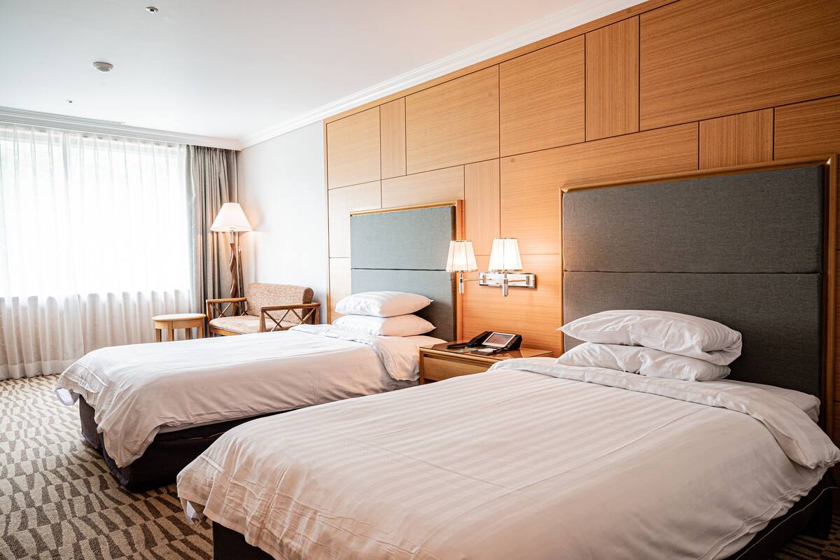 A hotel room with two beds, a wooden headboard, a small table with a lamp, and a chair.