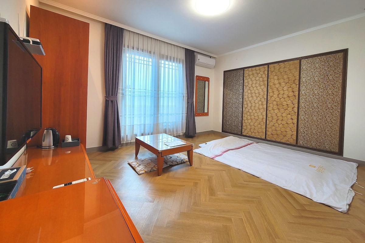 A hotel room with a wooden desk, a bed with a white comforter, a large window with sheer curtains, a wall-mounted air conditioner, and a traditional sliding door.