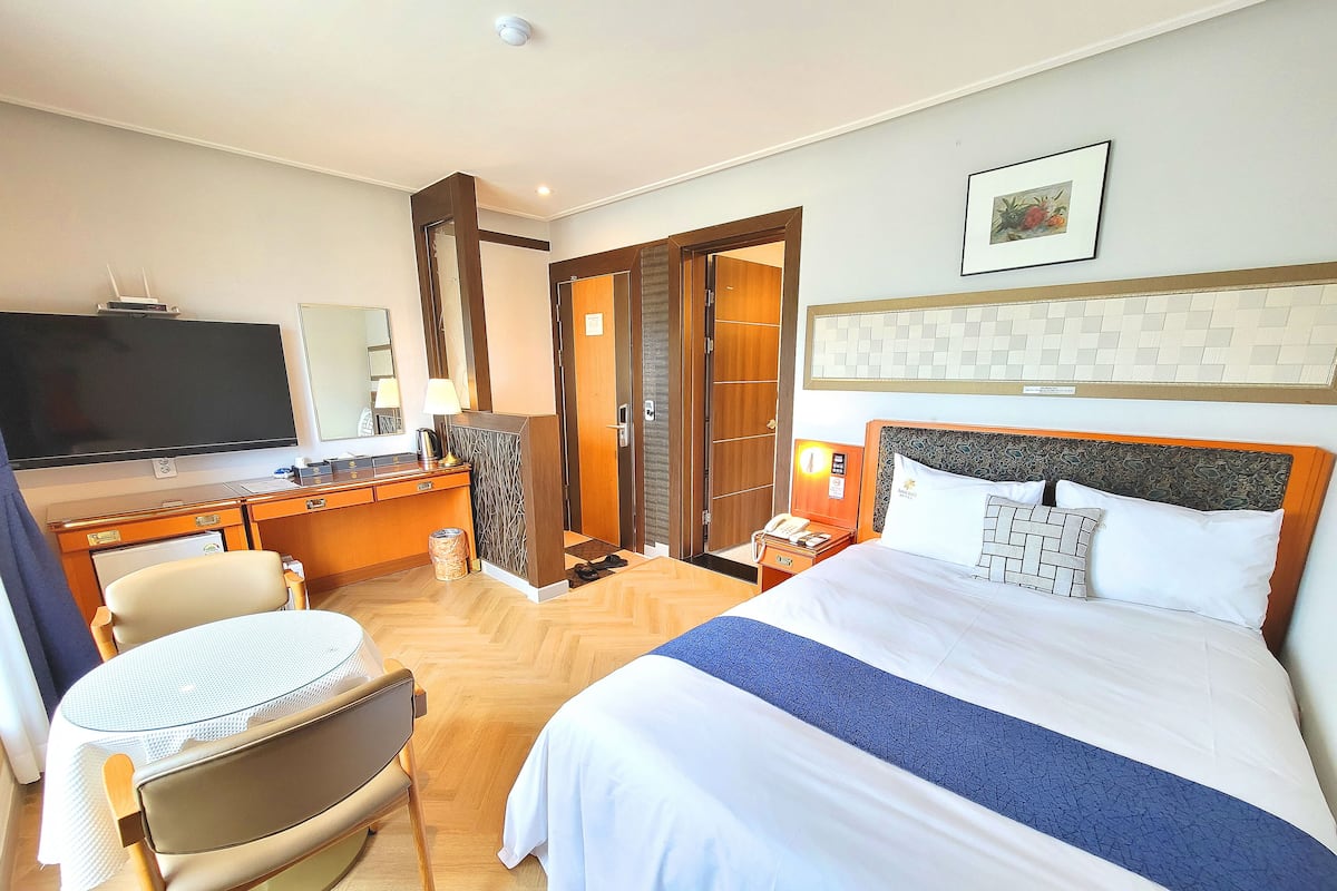 A hotel room with a bed, a desk with a computer, a chair, a television, and a door leading to another room.