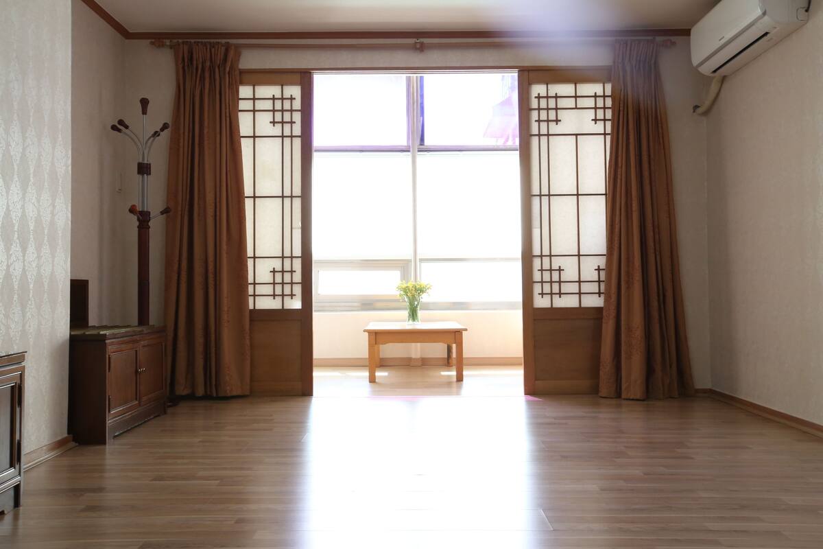 Ein Zimmer mit Holzboden, einem Holztisch mit einer Vase und einem Fenster mit braunen Vorhängen.
