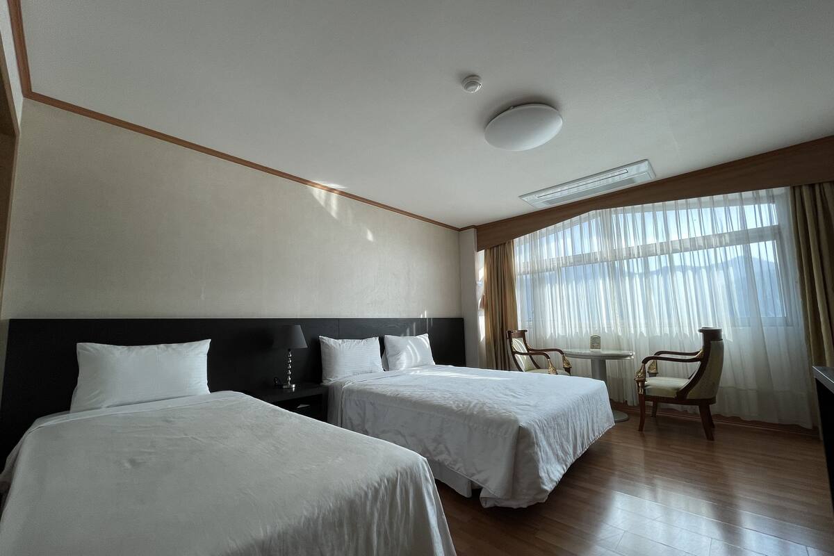 Ein Hotelzimmer mit zwei Betten, einem kleinen Tisch, einem Stuhl und einem großen Fenster mit Vorhängen.