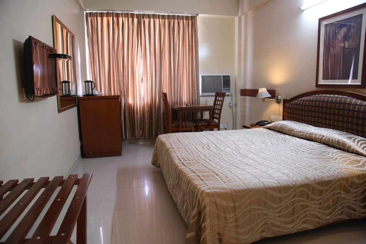 A hotel room with a bed, a desk, a chair, a wardrobe, a television, and an air conditioning unit.