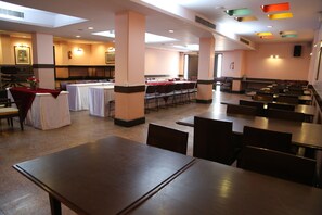 A spacious dining area with multiple tables and chairs, a long counter with a red tablecloth, and a kitchen area in the background.