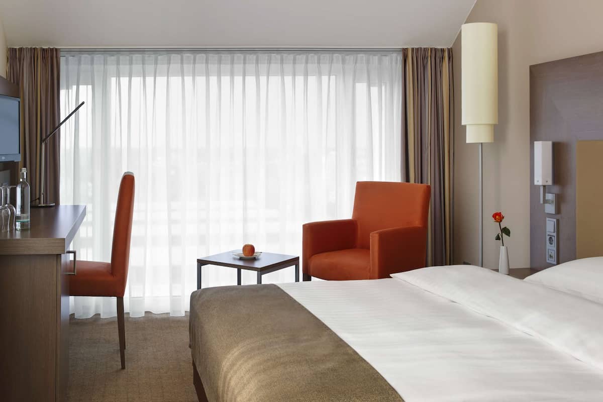 A hotel room with a bed, a desk with a chair, a small table with a fruit, and a lamp.