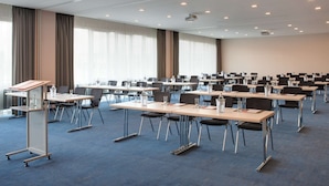 Un salón de conferencias con filas de mesas y sillas, amplias ventanas con cortinas y proyectores en el techo.