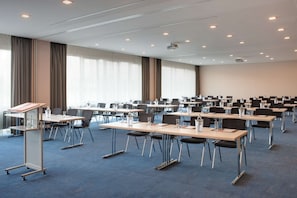 Un salón de conferencias con filas de mesas y sillas, amplias ventanas con cortinas y proyectores en el techo.