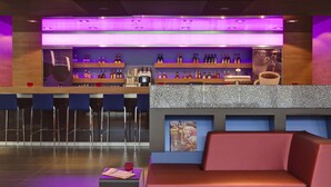 Zona de bar con techo iluminado en morado, barra con múltiples estantes de botellas y área de asientos con cojines rojos.