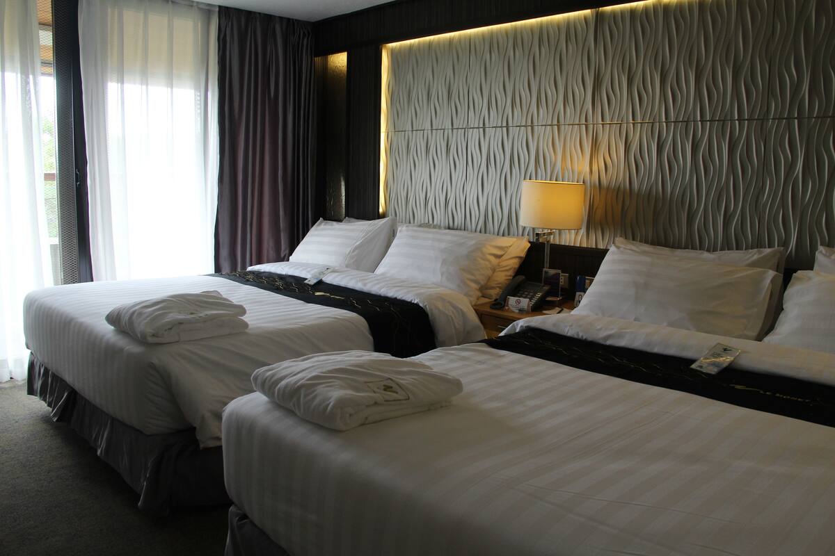 A hotel room with two beds, a headboard with a textured pattern, a bedside lamp, and a window with curtains.