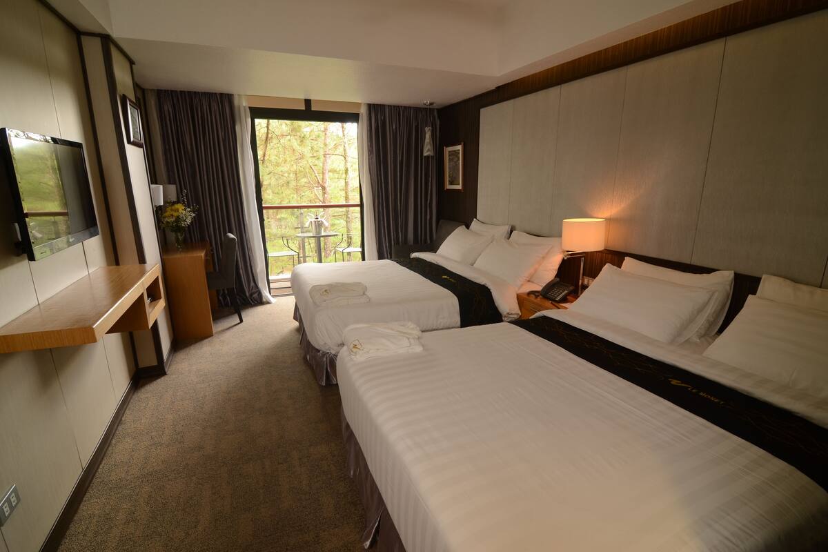 A hotel room with two beds, a TV, a desk, and a balcony with curtains.