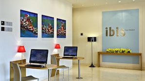 A modern hotel lobby with two desks, two chairs, a computer, a lamp, and a wall with the hotel name. - ibis Navi Mumbai Hotel (Navi Mumbai)