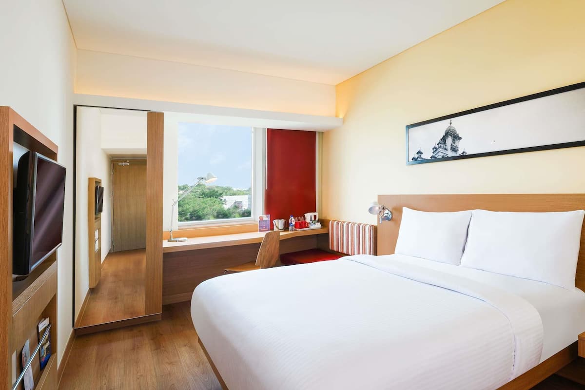 A hotel room with a bed, a desk, a TV, a window with a view, and a painting on the wall.