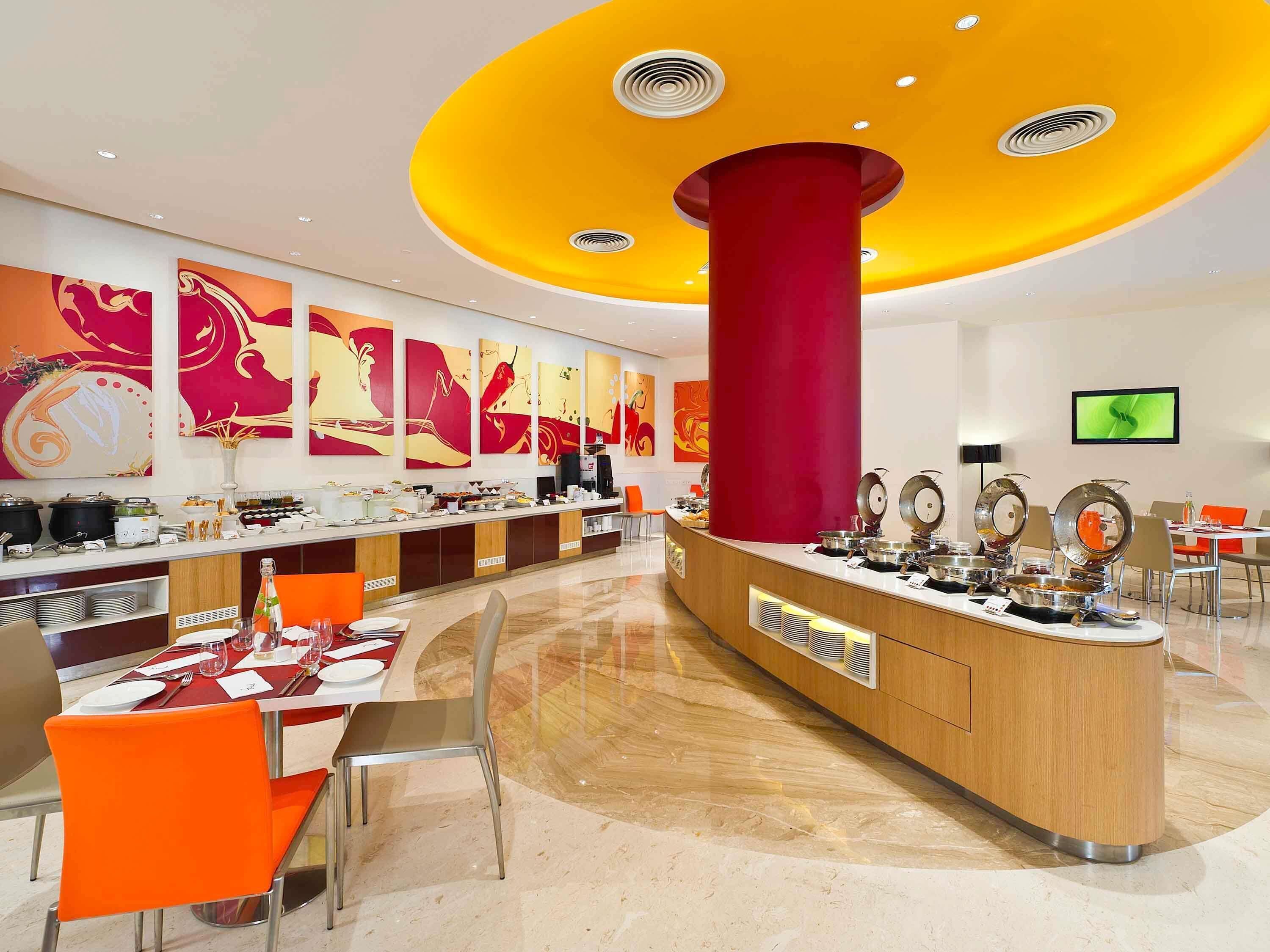A modern hotel lobby with a central reception desk, a buffet area, and a seating area with orange chairs.