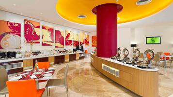 A modern hotel lobby with a central reception desk, a buffet area, and a seating area with orange chairs.