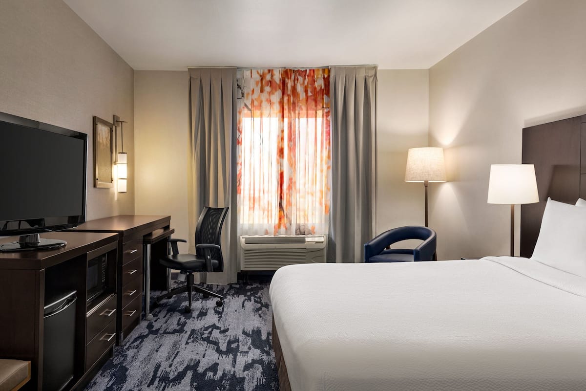 A hotel room with a bed, a desk with a television, a chair, a lamp, and a window with curtains.