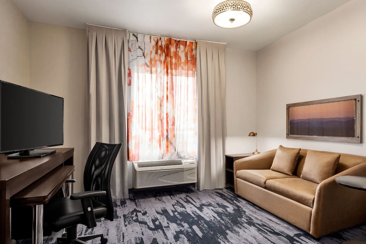A hotel room with a flat-screen TV, a leather sofa, a desk with a chair, a large window with curtains, and a painting on the wall.