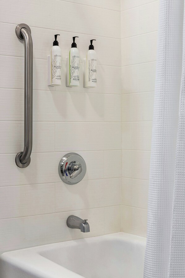 Un baño con una bañera blanca, un pasamanos y tres botellas de champú Alice & Co. en la pared.