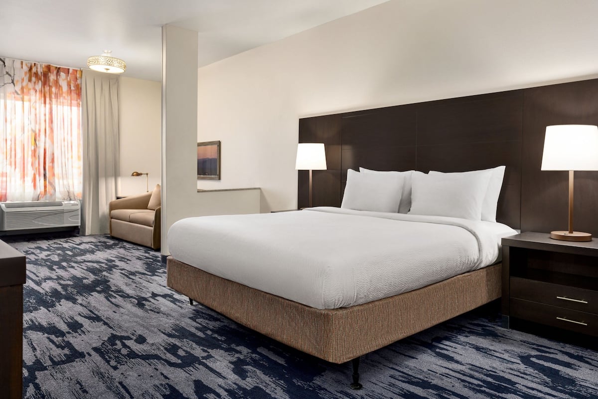 A hotel room with a large bed, two bedside tables with lamps, a chair, a desk, and a large window with curtains.