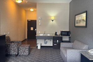 Habitación de hotel con kitchenette, un sofá, una silla, un cesto de basura, microondas y un cuadro en la pared.