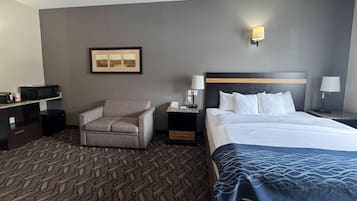 Una camera d'albergo con un letto, una sedia, una scrivania con un computer, un comodino con una lampada e un quadro appeso al muro.