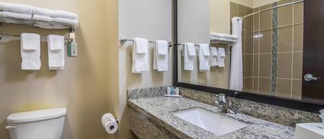 Un baño con inodoro, lavamanos y espejo. Hay toallas colgadas en un perchero y un dispensador de toallas.
