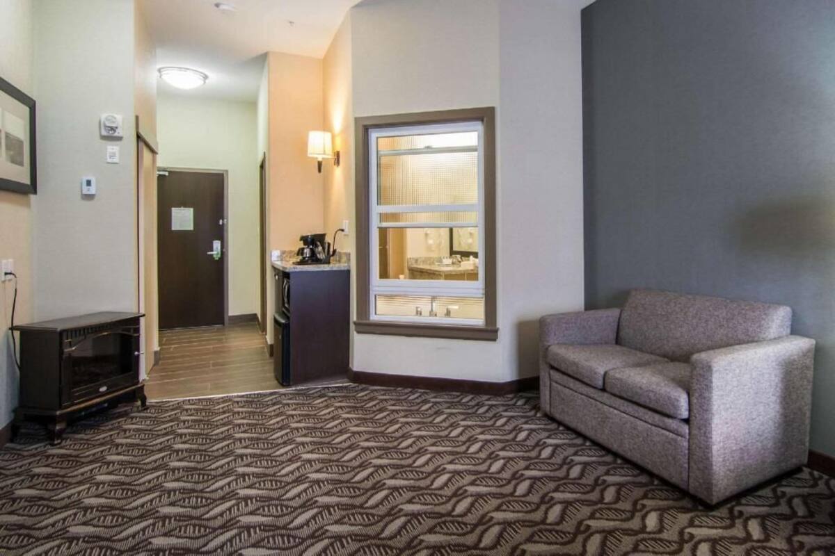 A hotel room with a patterned carpet, a sofa, a coffee maker, a window with blinds, and a door leading to another room.