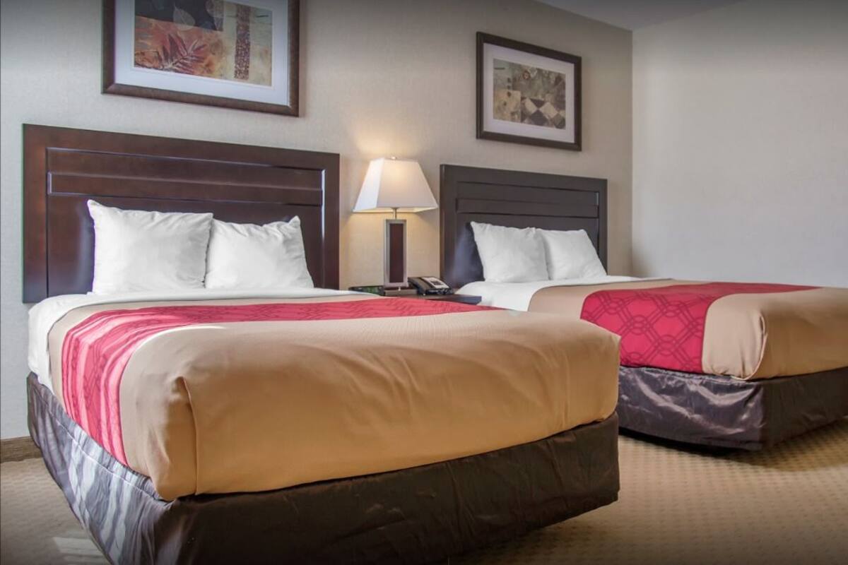 A hotel room with two beds, each with a wooden headboard, white pillows, and red and beige bedding. There is a nightstand with a lamp between the beds.