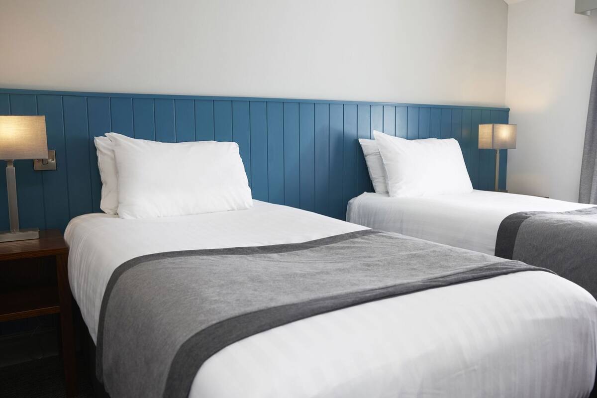 A hotel room with two beds, a blue headboard, bedside lamps, and a window with curtains.