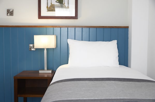 Una cama bien hecha con un almohadón blanco y una manta gris, una mesita de noche con una lámpara, y un cuadro en la pared.