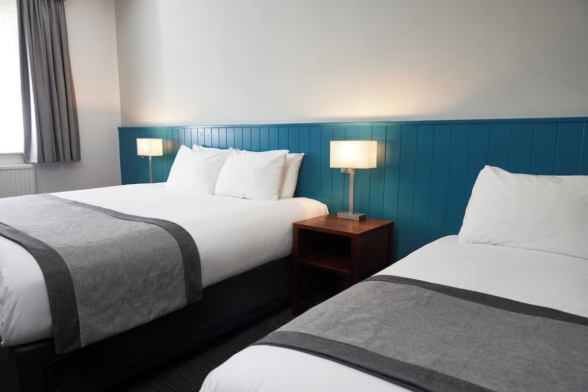 A hotel room with two beds, each with white bedding and a grey accent. There are two bedside tables with lamps, and the walls are painted teal.