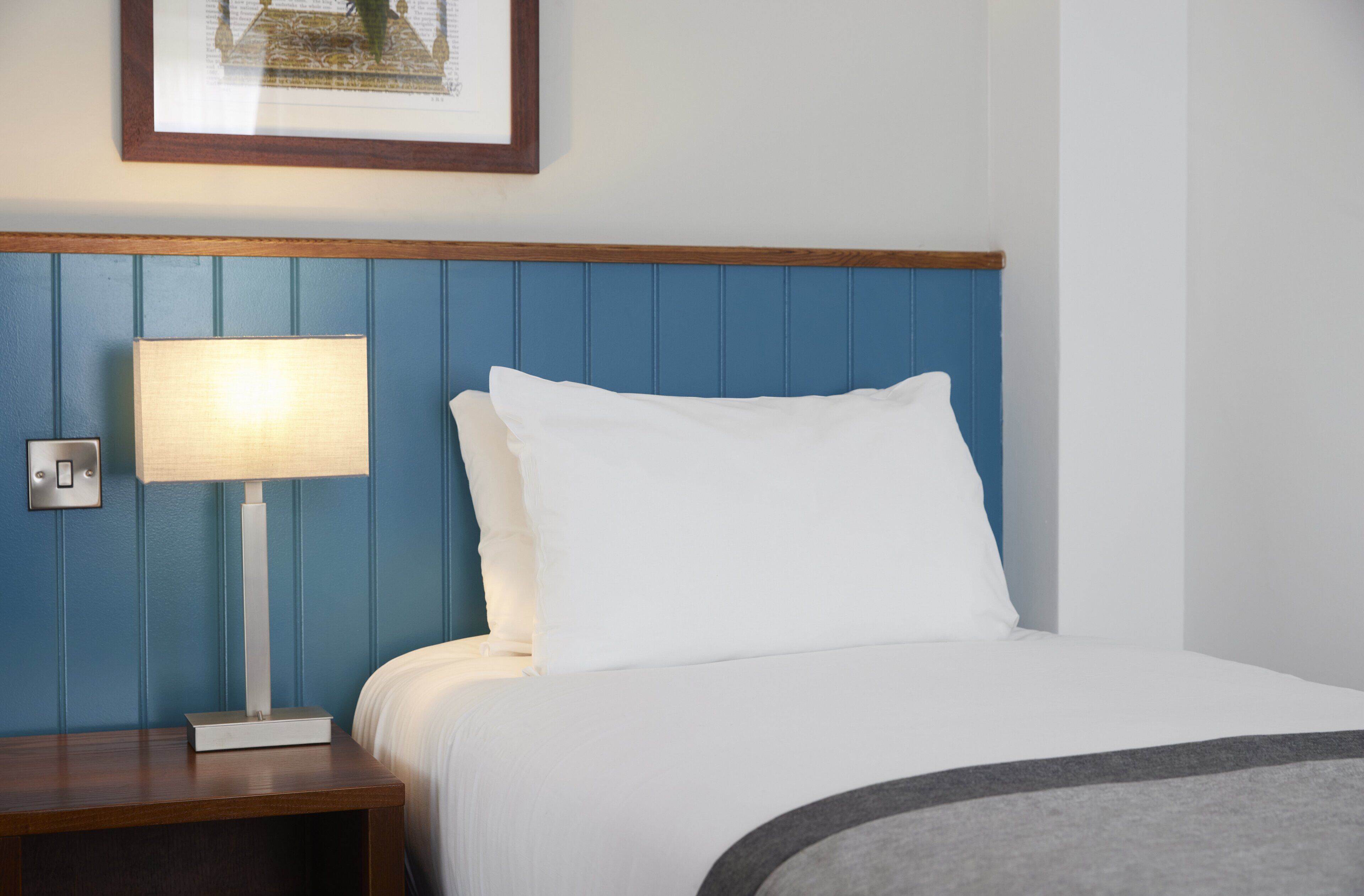 Una cama bien hecha con sábanas blancas y un plaid gris, una lámpara de noche y un cuadro en la pared.