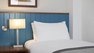 Una cama bien hecha con sábanas blancas y un plaid gris, una lámpara de noche y un cuadro en la pared.