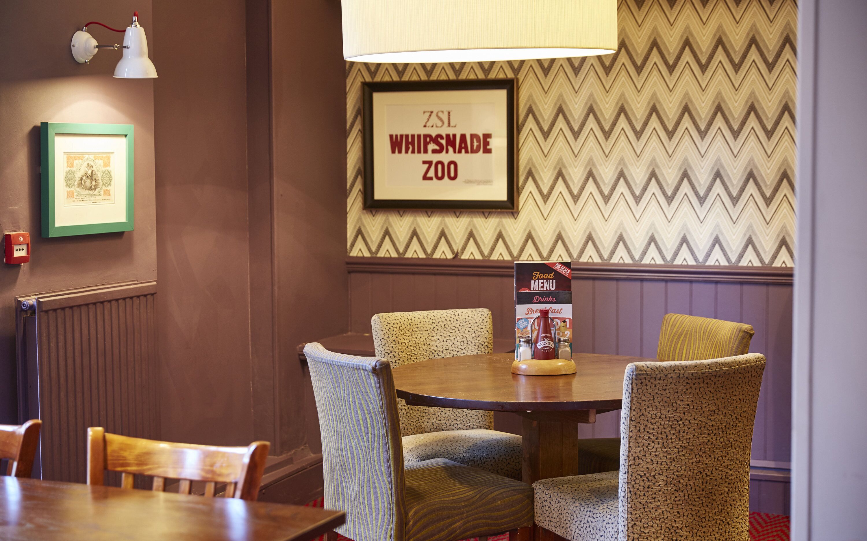 Un comedor con una mesa redonda, cuatro sillas tapizadas, un cartel enmarcado del Zoológico de ZSL Whipsnade, un cuadro enmarcado y un atril para el menú.