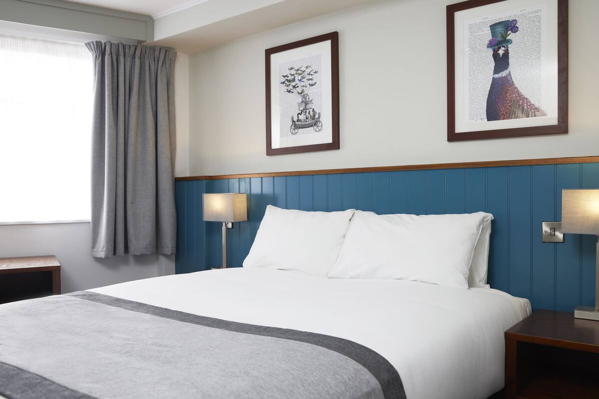 A neatly made bed with white linens and a grey accent, flanked by two bedside tables with lamps, in a room with a blue paneled headboard and two framed artworks on the wall.