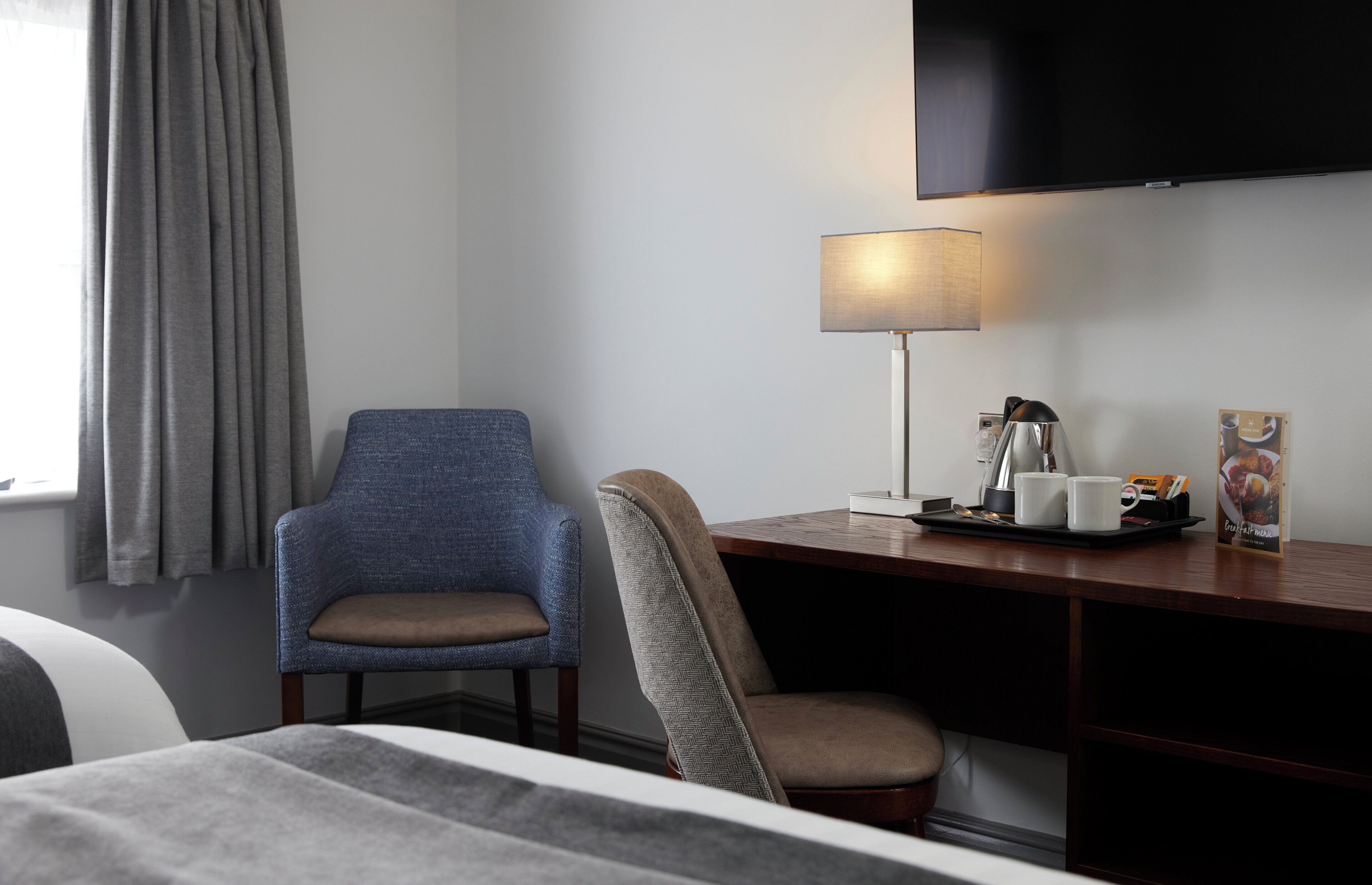 Quarto de hotel com cama, duas cadeiras, escrivaninha com luminária, televisão de tela plana e janela com cortinas.