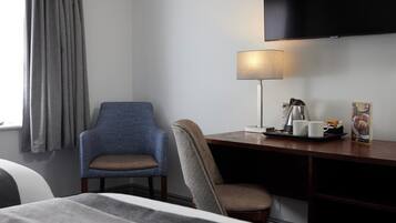 Quarto de hotel com cama, duas cadeiras, escrivaninha com luminária, televisão de tela plana e janela com cortinas.