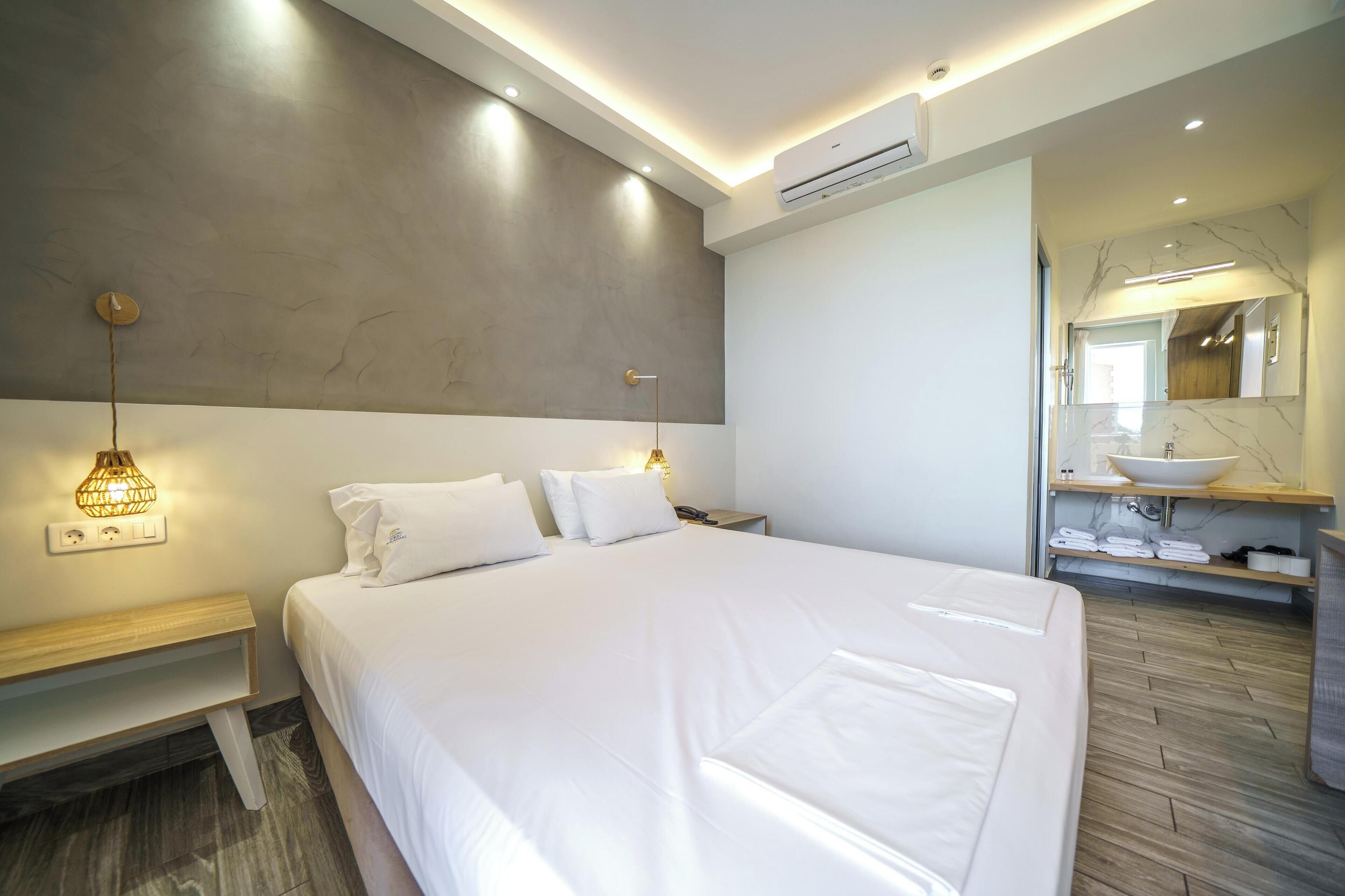 A modern hotel room with a large bed, bedside tables, a nightstand, a wall-mounted lamp, a built-in shelf with towels, and a bathroom with a vanity and mirror.