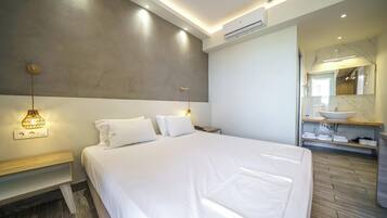A modern hotel room with a large bed, bedside tables, a nightstand, a wall-mounted lamp, a built-in shelf with towels, and a bathroom with a vanity and mirror.
