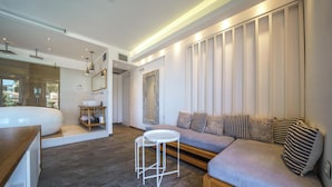 A modern living room with a grey sofa, a small round table, and a wooden bench. There is a large mirror on the wall and a window with blinds. - Porto Kalamaki Hotel (Chania)