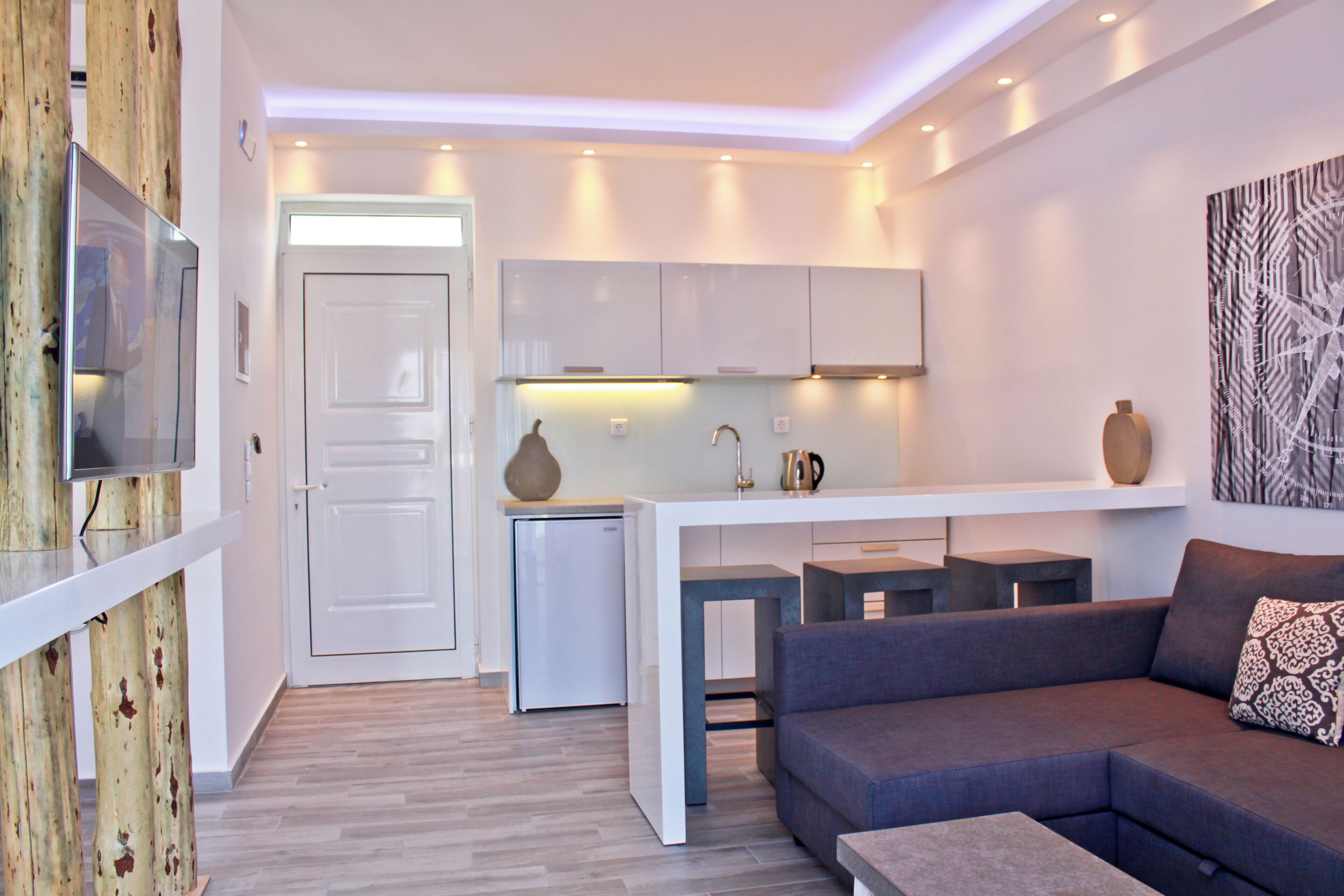 A modern living room with a grey sofa, a coffee table, and a TV mounted on a wooden wall. A kitchen area with a white fridge and cabinets is visible in the background.
