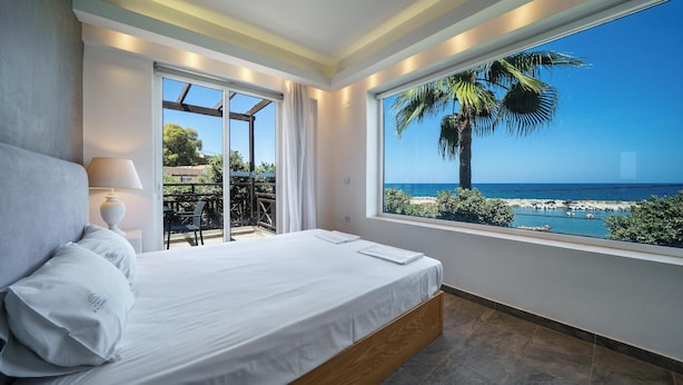 A bedroom with a large window overlooking a beach and palm tree, a bed with white linens, and a view of the sea. - Porto Kalamaki Hotel (Chania)