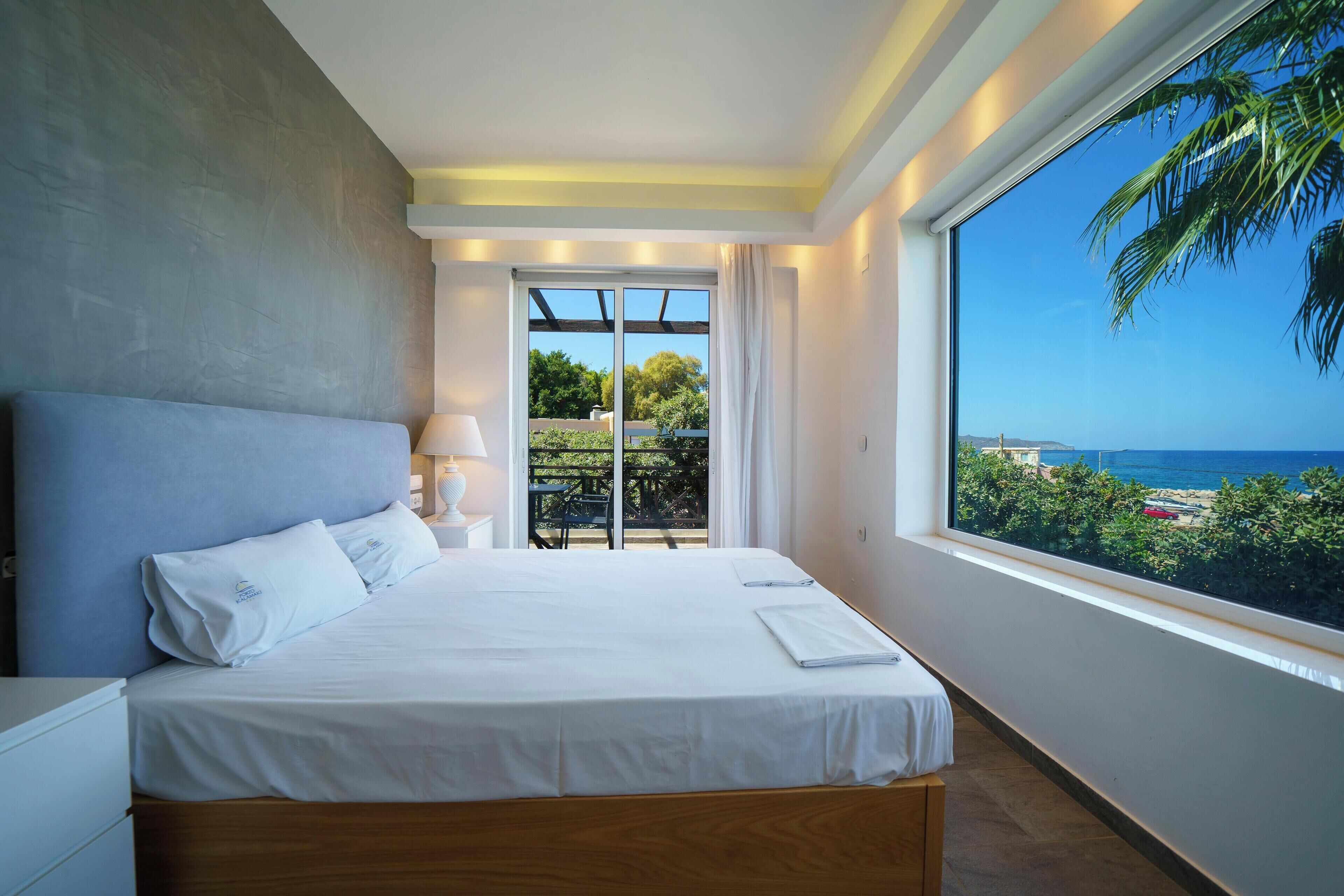 A modern hotel room with a large bed, a view of the sea, and a balcony with outdoor furniture.