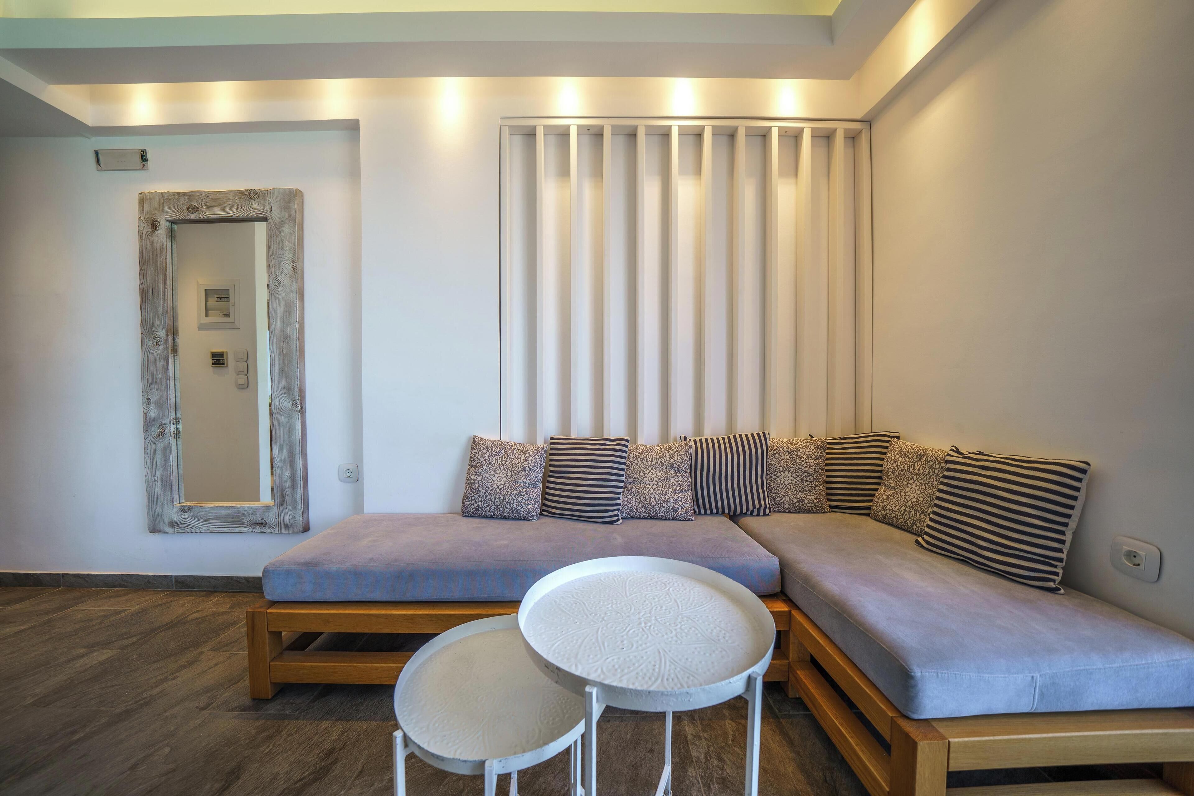 Una sala moderna con un sofá modular, dos mesas redondas blancas y un amplio ventanal con persianas verticales.