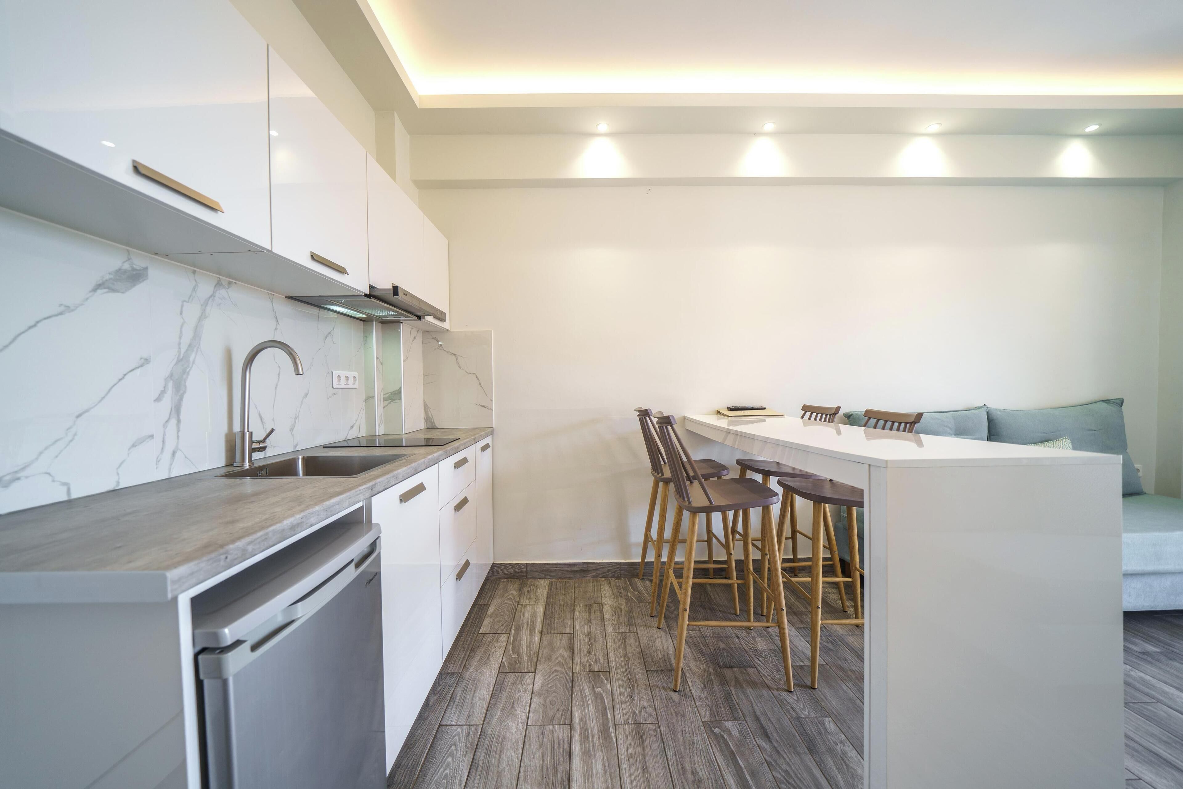 A modern kitchen with white cabinets, a marble backsplash, and a wooden countertop. There is a built-in dishwasher and a bar area with stools.