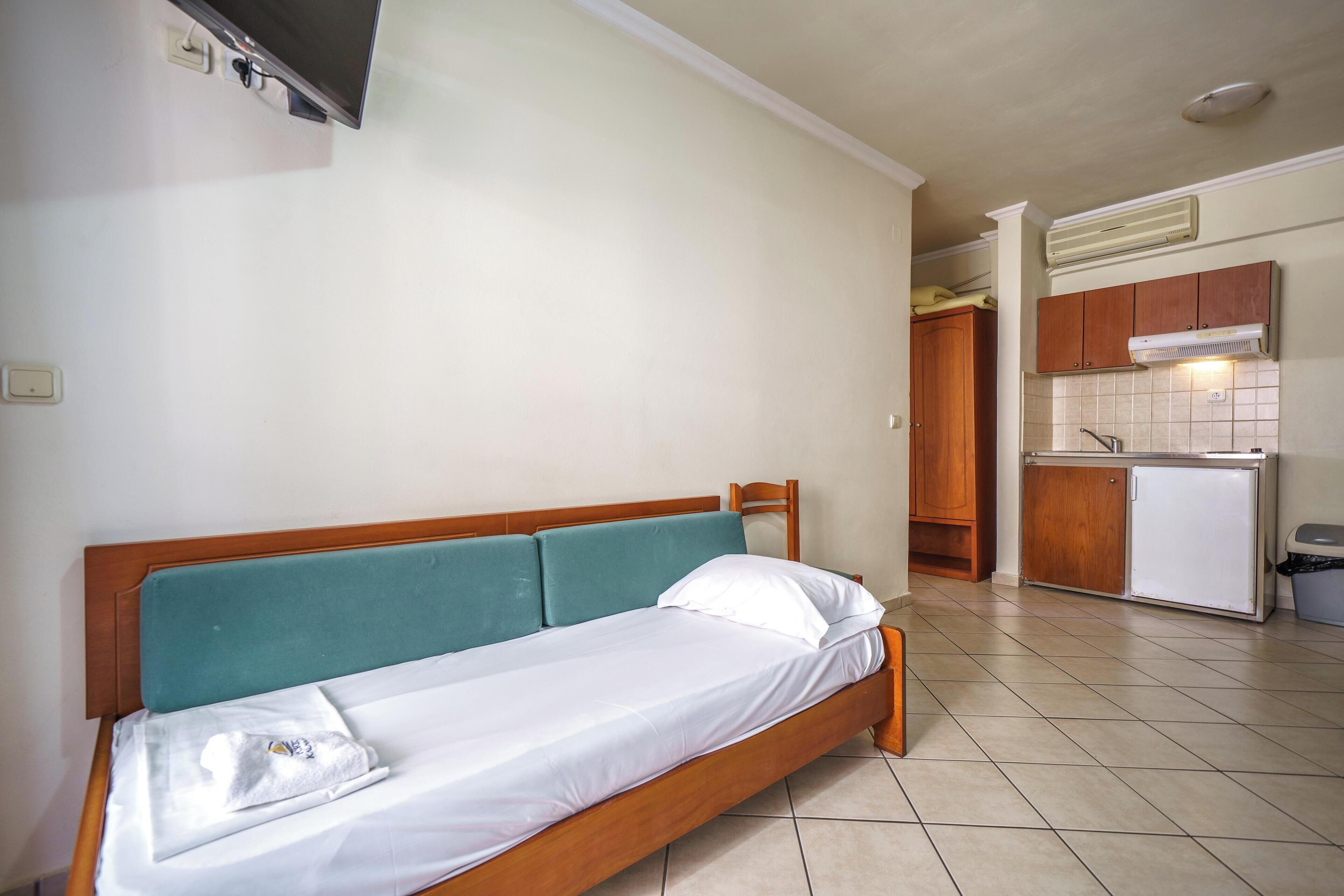 A single bed with a wooden frame and teal headboard, a small kitchenette with a refrigerator and wooden cabinets, and a tiled floor.