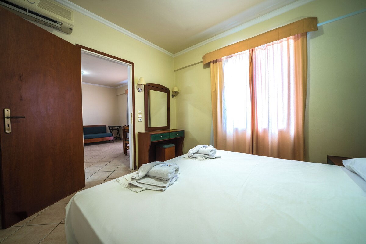 A hotel room with a bed, a wooden dresser, a mirror, and a door leading to another room.