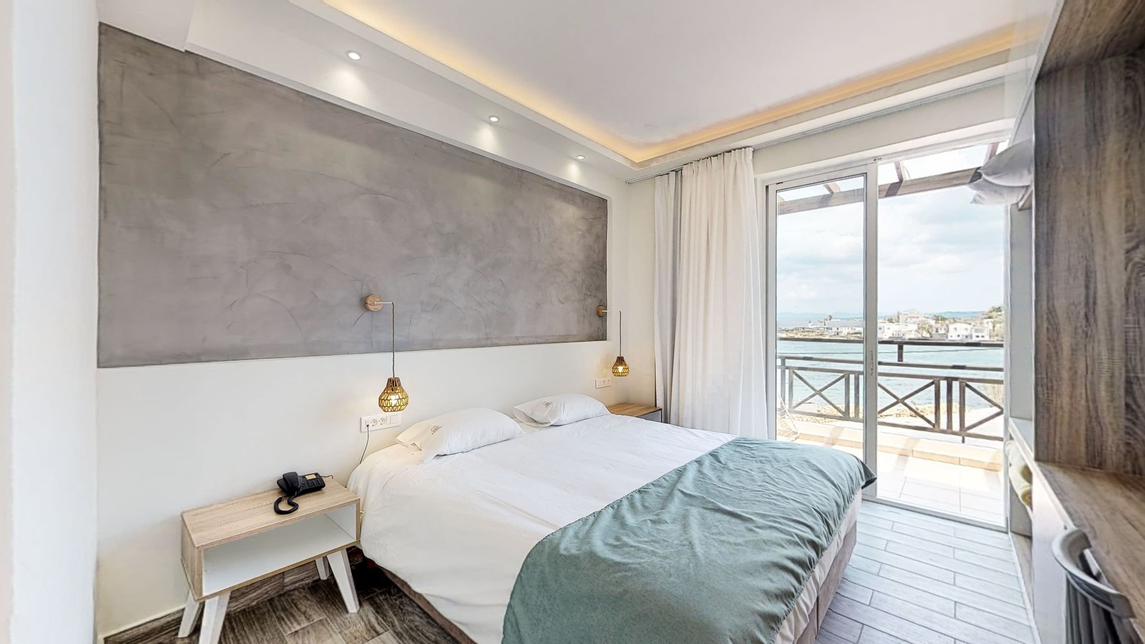 Habitación de hotel moderna con una cama grande, lámparas de noche, un teléfono y vistas al mar desde un balcón.