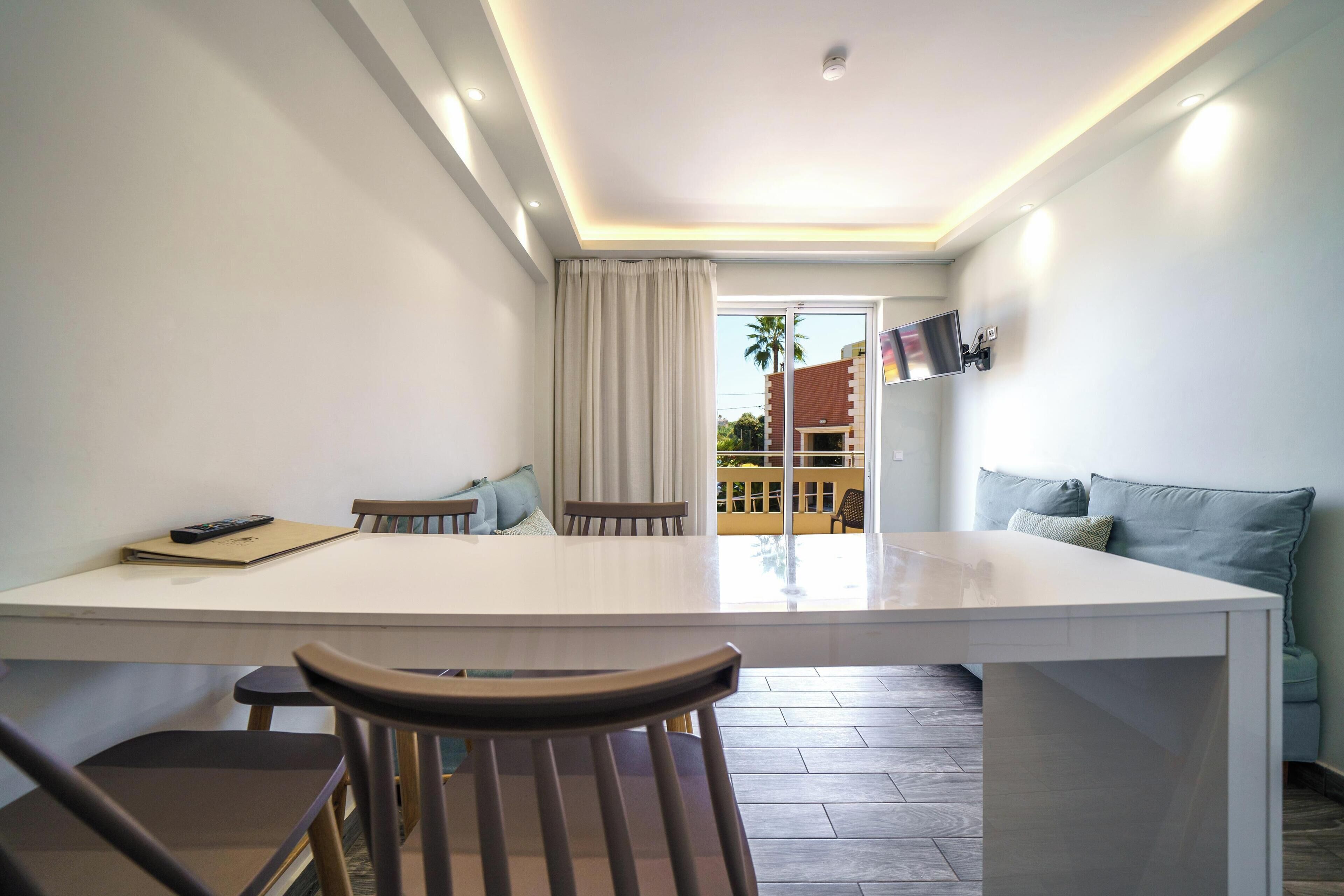 A modern living room with a large white table, two wooden chairs, a sofa with cushions, a TV mounted on the wall, and a view of the outside through a glass door.