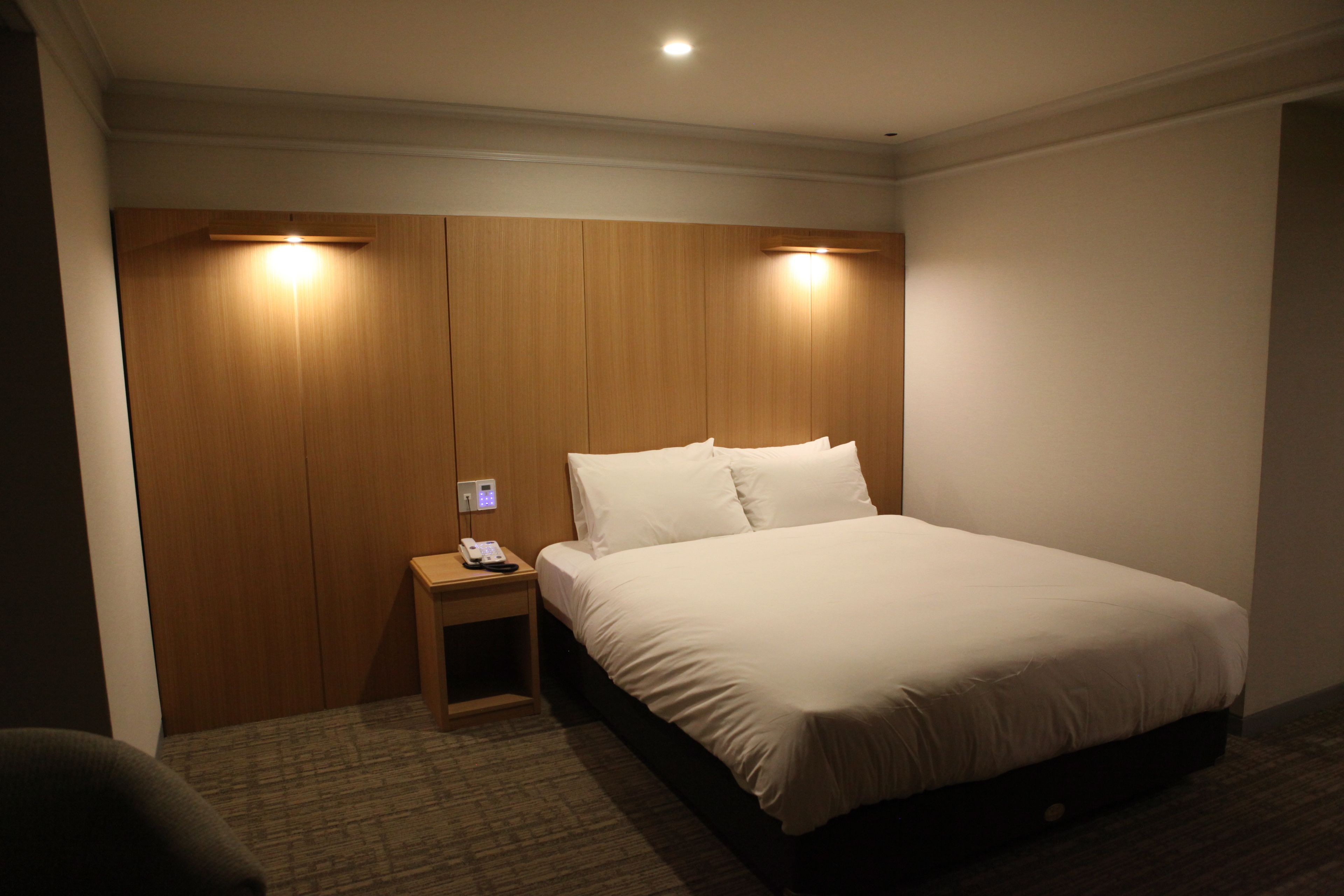 A hotel room with a wooden headboard, a single bed with white bedding, a bedside table with a phone, and a carpeted floor.