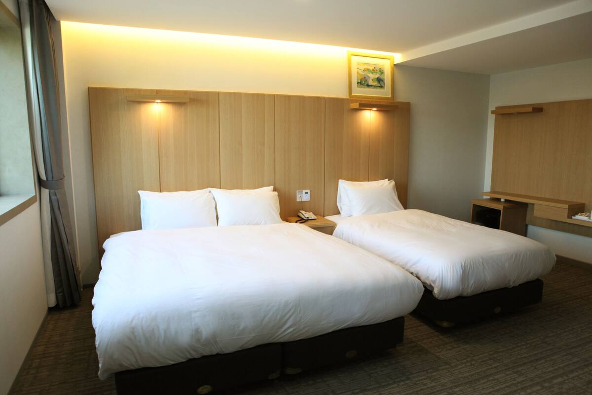 A hotel room with two beds, a wooden headboard, a bedside table with a lamp, and a small shelf.