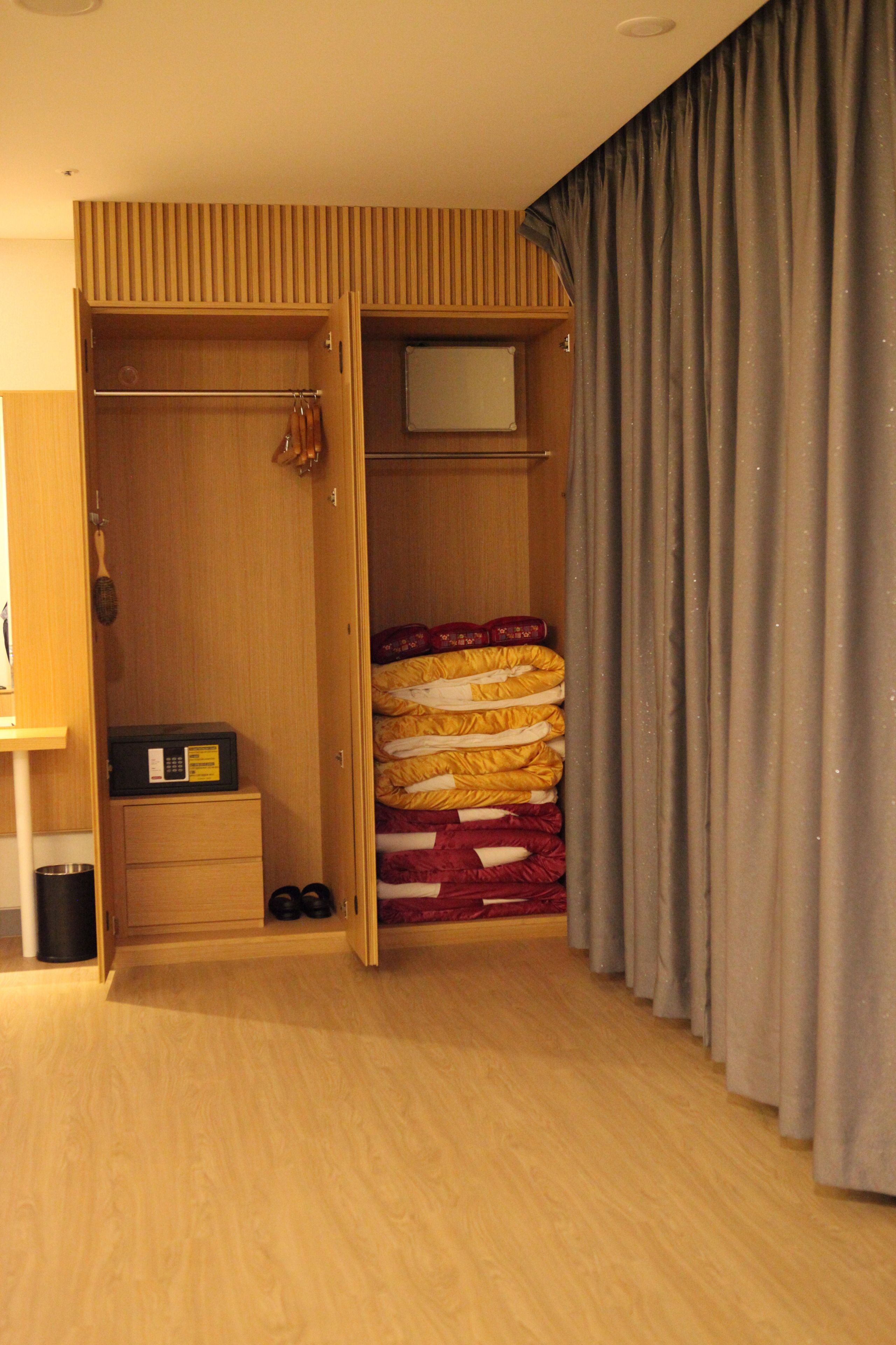 A bedroom with a wooden wardrobe, a bed with a red and yellow blanket, and grey curtains.